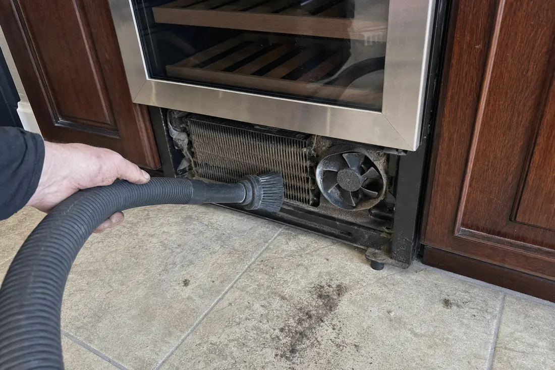 A brush and vacuum hose on the dust-loaded condenser coil behind a removed grille - Sub-Zero refrigeration repair
