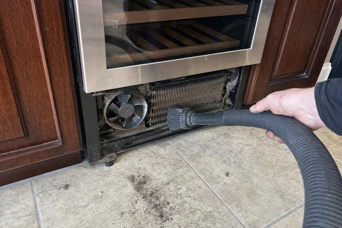 A brush and vacuum hose on the dust-loaded condenser coil behind a removed grille, the matted coil fins in frame - Built-in refrigeration repair