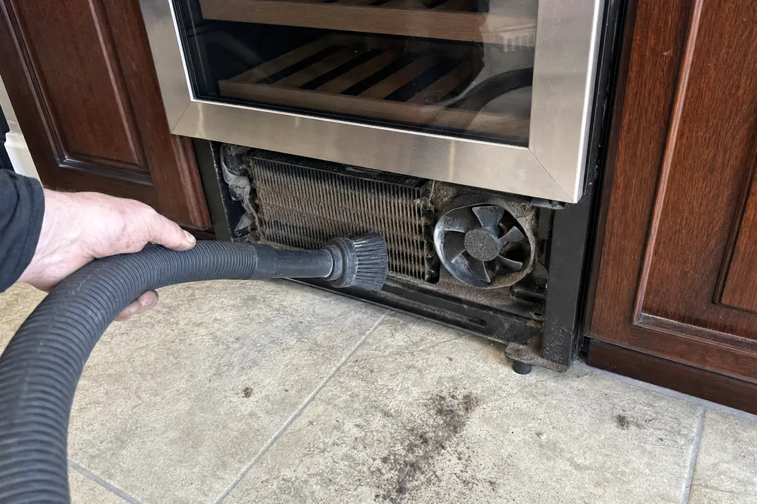 A brush and vacuum hose on the dust-loaded condenser coil behind a removed grille, the vacuum hose in frame - Equipment we repair