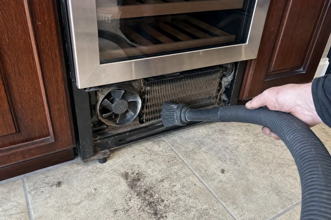 A brush and vacuum hose on the dust-loaded condenser coil behind a removed grille, the soft brush in frame - What a visit costs
