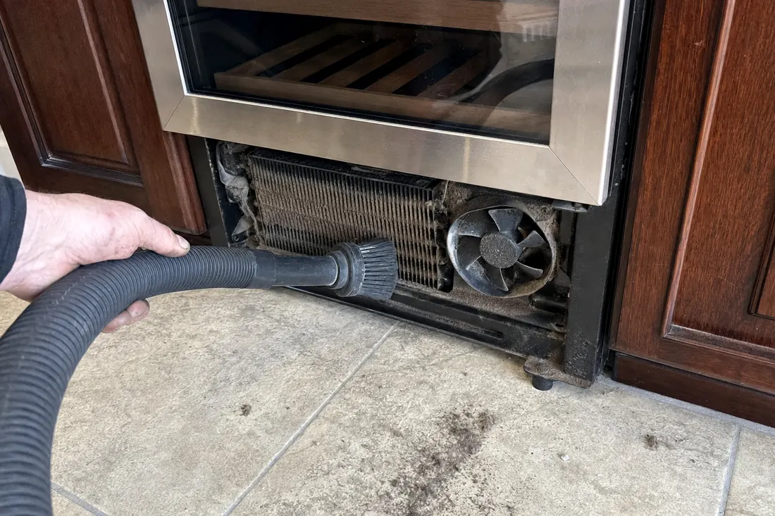 A brush and vacuum hose on the dust-loaded condenser coil behind a removed grille, dust on the floor tile in frame - Privacy notice
