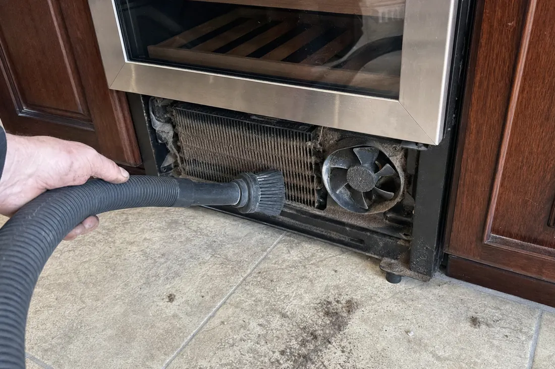 A brush and vacuum hose on the dust-loaded condenser coil behind a removed grille, the matted coil fins visible in the picture - What the repair is warranted