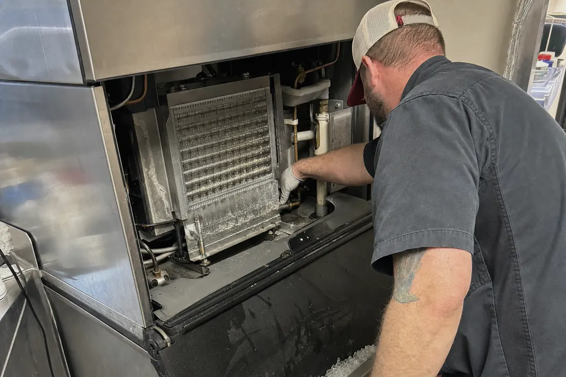 Technician at a stainless ice machine with the top panel off, checking the water line and the evaporator plate - Accessibility