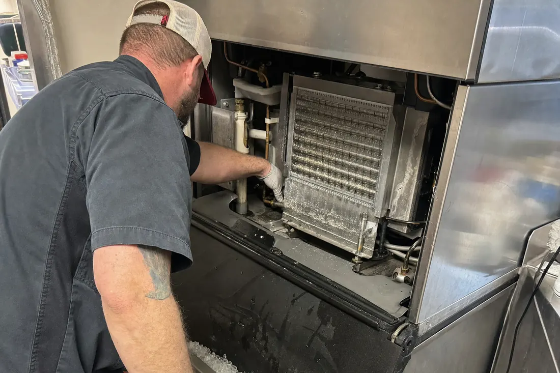 Technician at a stainless ice machine with the top panel off, checking the water line and the evaporator plate, the evaporator plate in frame - Book a visit