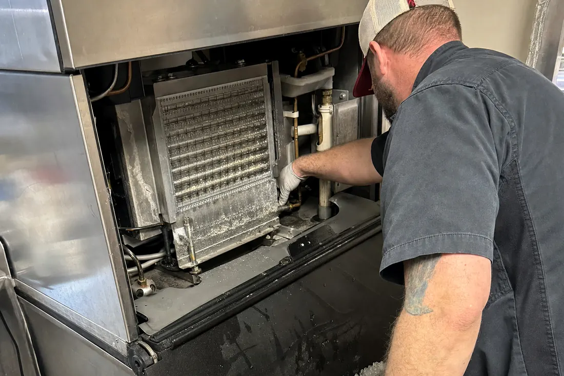 Technician at a stainless ice machine with the top panel off, checking the water line and the evaporator plate, the removed top panel in frame - Appliance repair in Jacksonville