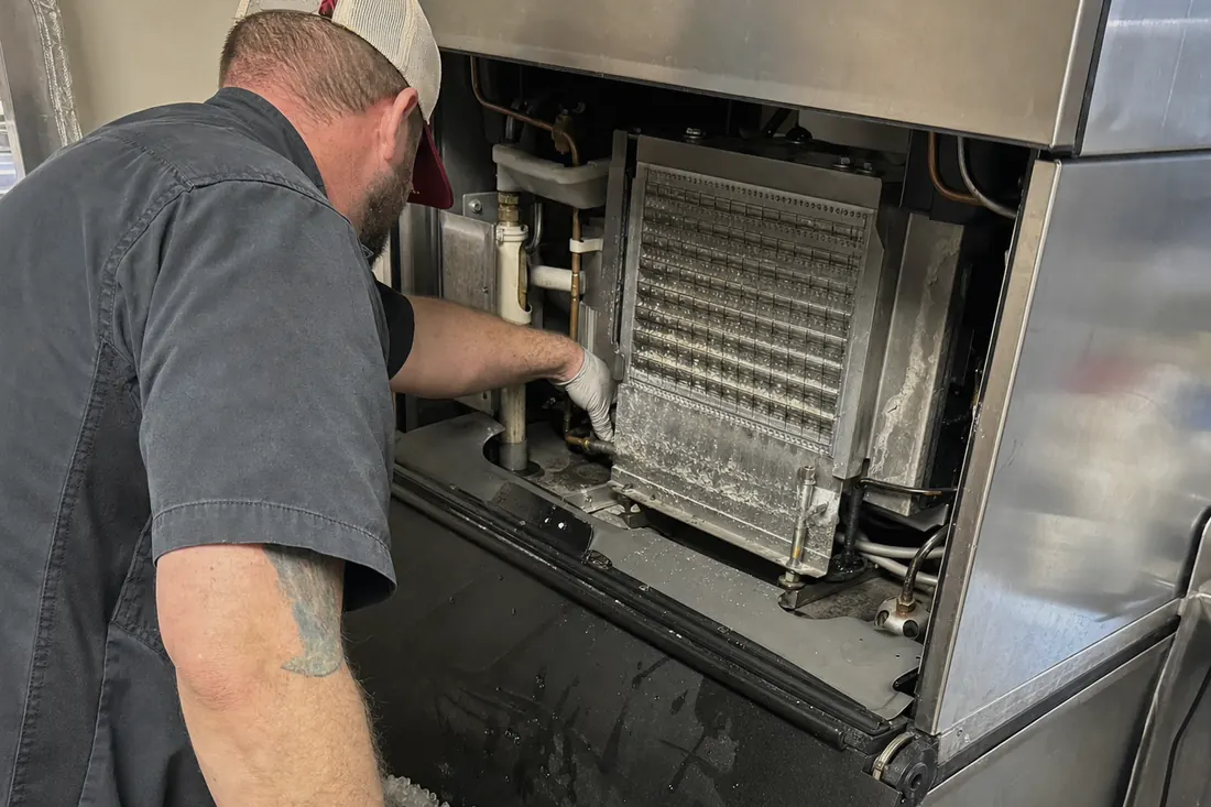 Technician at a stainless ice machine with the top panel off, checking the water line and the evaporator plate, the ice bin in frame - Hurricane outages