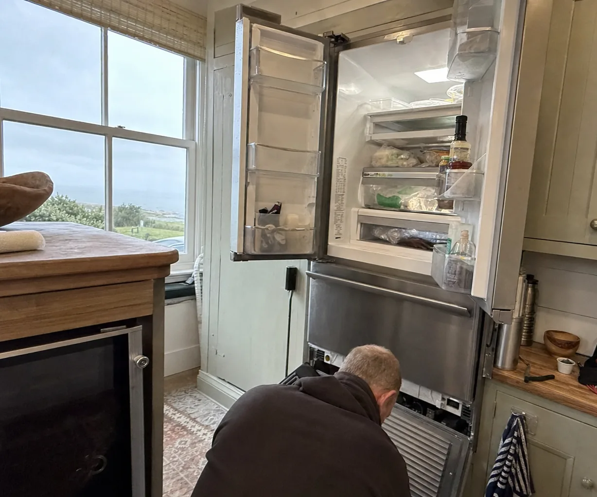 Technician kneeling at the open lower grille of a built-in French-door refrigerator with a freezer drawer, a window over the sea beside him