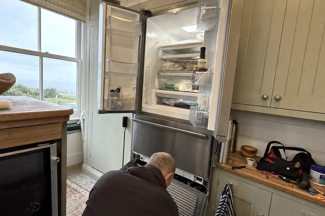 An open built-in refrigerator in a coastal kitchen, technician at the lower grille, the open doors in frame - Where we work