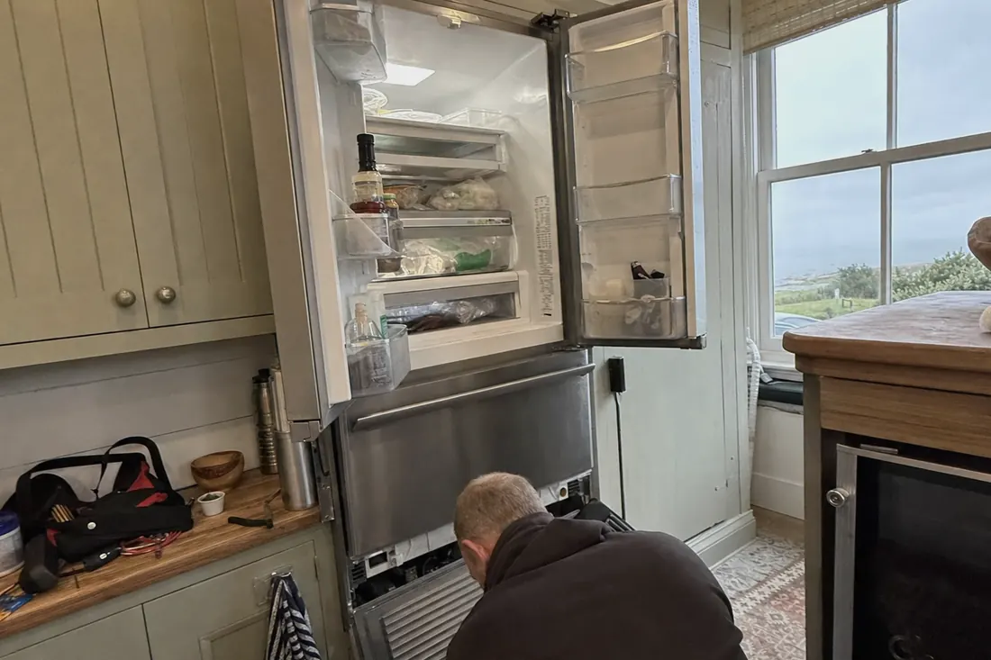An open built-in refrigerator in a coastal kitchen, technician at the lower grille, the daylight at the window in frame - Appliance repair in St. Augustine and St. Johns